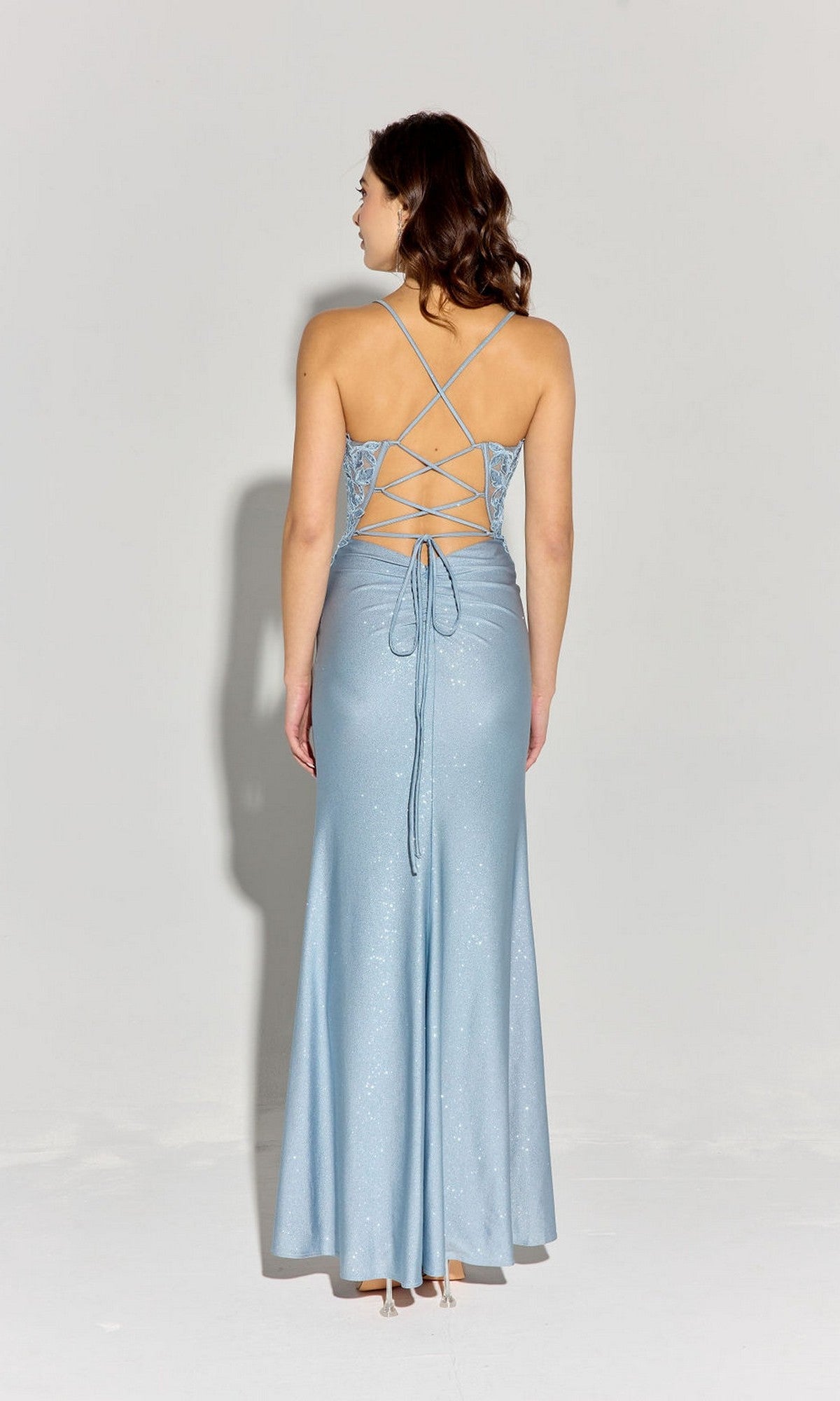 A woman faces away from the camera wearing the Eureka EK129, a light blue, sleeveless long prom dress featuring a glitter jersey sheath skirt and lace-up open back, set against a plain background.